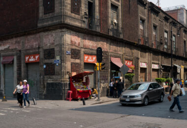 A busy corner in Mexico City’s Centro Histórico where Calle República de Uruguay meets Calle 5 de Febrero. The block’s centuries-old volcanic stone buildings, typical of the colonial period, now house small shops and street vendors that reflect the area’s dense commercial activity. The facade’s weathered cantera stone and red tezontle give the structure its distinctive character, common in pre-19th century civic architecture throughout the historic core.