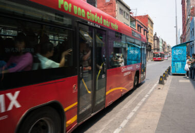 A Mexico City Metrobus on Line 4 passes through the narrow streets of the Centro Histórico, heading toward the San Lázaro terminus. This route, introduced in 2012, was designed to connect the downtown core with the city’s main transit hubs and the airport while reducing congestion in one of Latin America’s most crowded historic districts. The red articulated buses operate along corridors like República de Uruguay and República de El Salvador, part of an effort to modernize and green urban mobility.