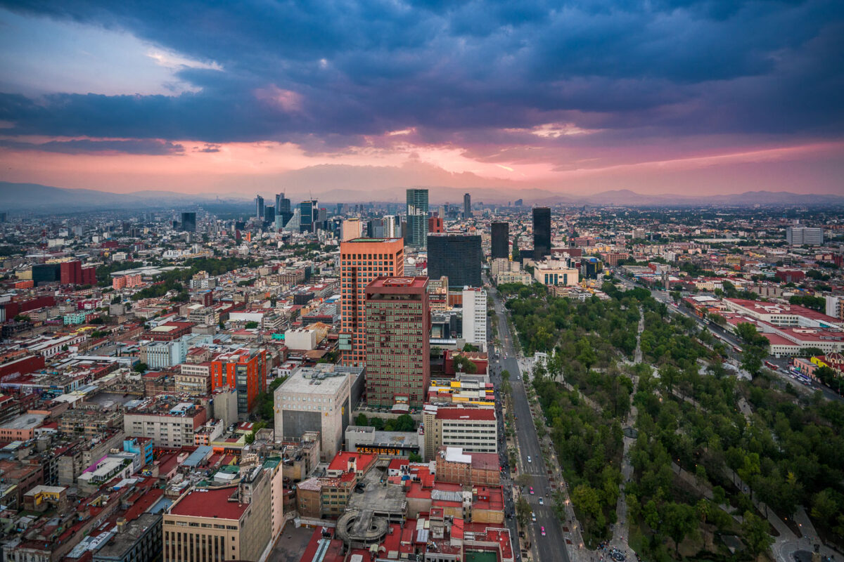 Mexico City is shown from above at sunset, with the Paseo de la Reforma running diagonally through the right side of the image. To the left, the dense central districts display mid-rise buildings with red rooftops, while clusters of high-rise office towers dominate the skyline further west. The large green area on the right is the Alameda Central park. In the distance, the Sierra Madre mountains form the backdrop under a layer of clouds.