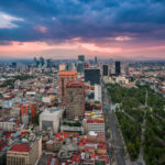 Mexico City is shown from above at sunset, with the Paseo de la Reforma running diagonally through the right side of the image. To the left, the dense central districts display mid-rise buildings with red rooftops, while clusters of high-rise office towers dominate the skyline further west. The large green area on the right is the Alameda Central park. In the distance, the Sierra Madre mountains form the backdrop under a layer of clouds.