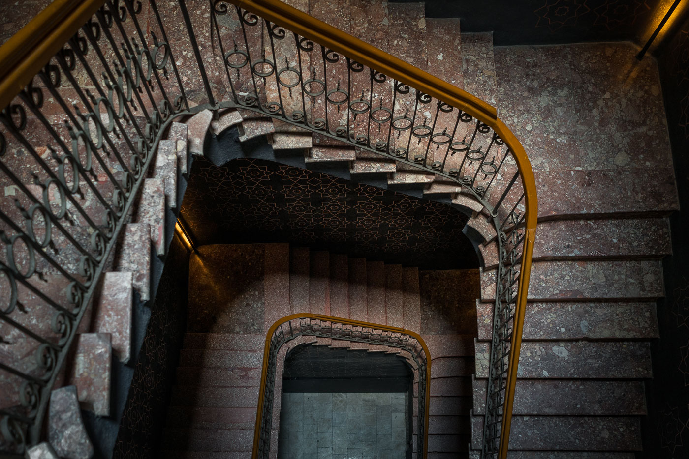 An intricately designed stairwell within a renovated Mexico City hotel, featuring red terrazzo stone steps and wrought-iron railings that spiral through multiple floors. The geometric patterns adorning the walls echo the Art Deco and neoclassical influences seen across the capital’s early 20th-century architecture. Thoughtful lighting accentuates the texture of the stonework, blending historic craftsmanship with the building’s modern restoration.