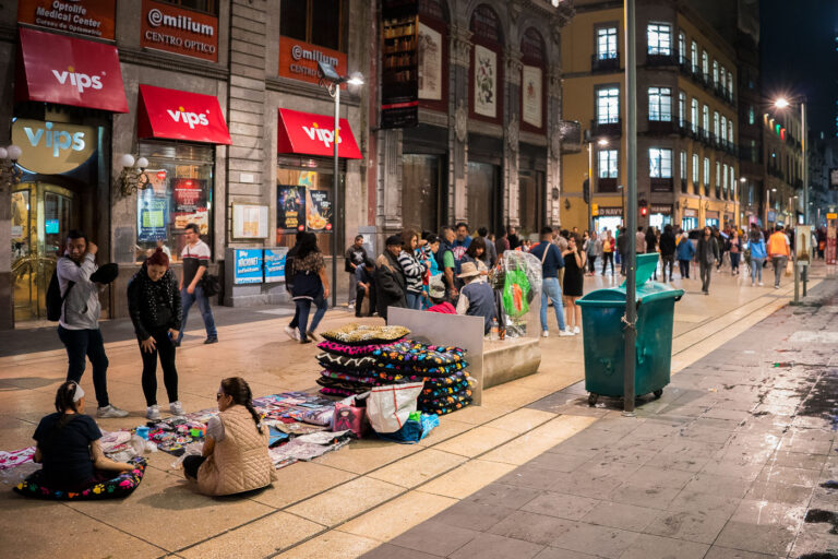 Street Vendors on Avenida Francisco I. Madero — Mexico City, 2 3 As evening settles over Mexico City’s Centro Histórico, informal vendors spread colorful textiles, pillows, and trinkets across the pedestrian walkway of Avenida Francisco I. Madero. The avenue, one of Latin America’s busiest foot corridors, connects the Zócalo to the Palacio de Bellas Artes and thrums with life well into the night. Illuminated storefronts and cafés like Vips cast reflections onto the restored stone façades of early 20th-century architecture. Beneath the glow of the streetlamps, the mingling of workers, families, and passersby captures the layered rhythm of Mexico City — a metropolis where commerce, tradition, and resilience coexist within its historic streetscape.