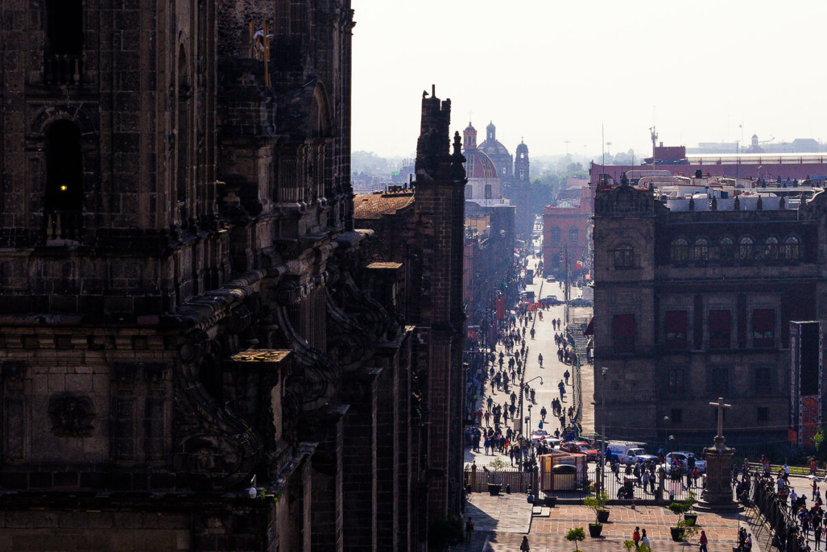 A busy street scene in Mexico City’s historic center, viewed from the steps of the Metropolitan Cathedral. Pedestrians fill the avenue leading eastward, lined with colonial-era buildings. In the background, the domes and towers of additional churches rise above the rooftops, marking the dense architectural landscape of the city’s downtown.