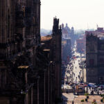 A busy street scene in Mexico City’s historic center, viewed from the steps of the Metropolitan Cathedral. Pedestrians fill the avenue leading eastward, lined with colonial-era buildings. In the background, the domes and towers of additional churches rise above the rooftops, marking the dense architectural landscape of the city’s downtown.