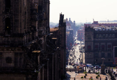 A busy street scene in Mexico City’s historic center, viewed from the steps of the Metropolitan Cathedral. Pedestrians fill the avenue leading eastward, lined with colonial-era buildings. In the background, the domes and towers of additional churches rise above the rooftops, marking the dense architectural landscape of the city’s downtown.