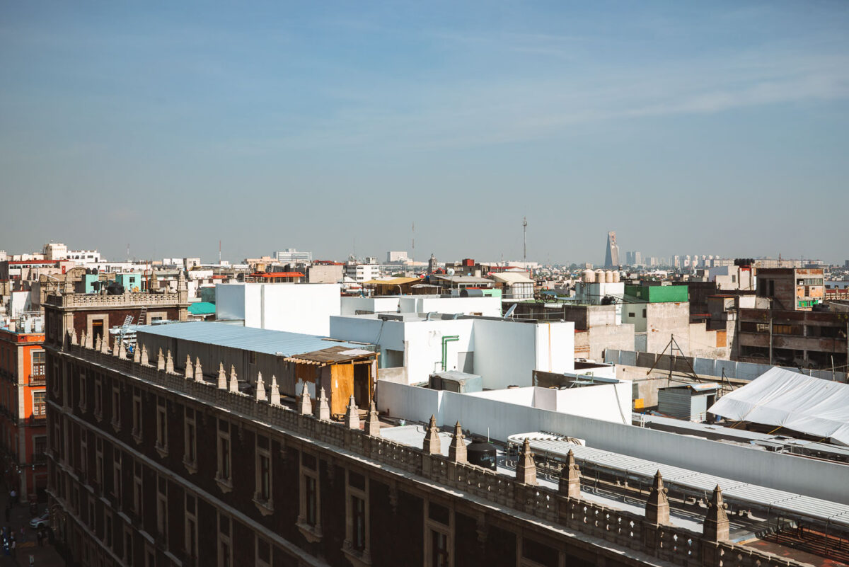 Rooftops of Mexico City’s historic center fill the foreground, with flat structures, water tanks, and makeshift additions visible across the skyline. Toward the horizon, modern high-rise buildings such as Torre Latinoamericana and Torre Reforma rise above the urban sprawl, contrasting with the older low-rise construction of the central district.