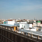 Rooftops of Mexico City’s historic center fill the foreground, with flat structures, water tanks, and makeshift additions visible across the skyline. Toward the horizon, modern high-rise buildings such as Torre Latinoamericana and Torre Reforma rise above the urban sprawl, contrasting with the older low-rise construction of the central district.