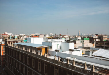 Rooftops of Mexico City’s historic center fill the foreground, with flat structures, water tanks, and makeshift additions visible across the skyline. Toward the horizon, modern high-rise buildings such as Torre Latinoamericana and Torre Reforma rise above the urban sprawl, contrasting with the older low-rise construction of the central district.