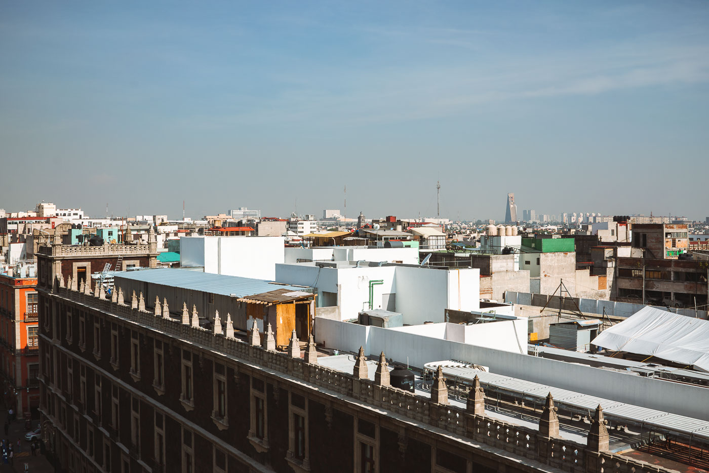 Rooftops of Mexico City’s historic center fill the foreground, with flat structures, water tanks, and makeshift additions visible across the skyline. Toward the horizon, modern high-rise buildings such as Torre Latinoamericana and Torre Reforma rise above the urban sprawl, contrasting with the older low-rise construction of the central district.