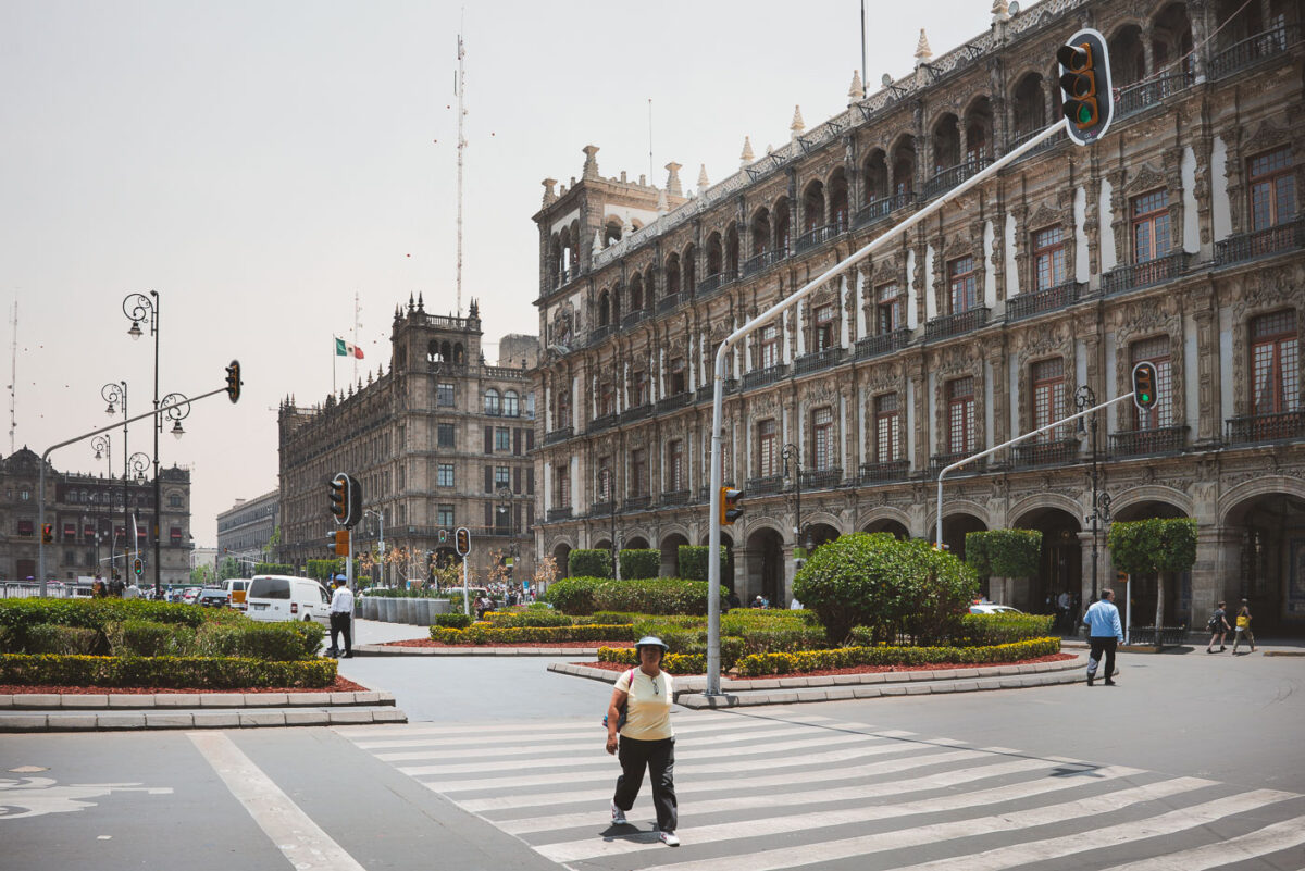 Pedestrians cross a street at Mexico City’s Zócalo, with the ornate colonial-era Government Palace and other historic administrative buildings lining the square. The Mexican flag is visible flying above the rooftops, while traffic lights and landscaped roundabouts frame the busy intersection.