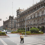Pedestrians cross a street at Mexico City’s Zócalo, with the ornate colonial-era Government Palace and other historic administrative buildings lining the square. The Mexican flag is visible flying above the rooftops, while traffic lights and landscaped roundabouts frame the busy intersection.