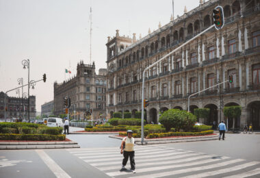 Pedestrians cross a street at Mexico City’s Zócalo, with the ornate colonial-era Government Palace and other historic administrative buildings lining the square. The Mexican flag is visible flying above the rooftops, while traffic lights and landscaped roundabouts frame the busy intersection.