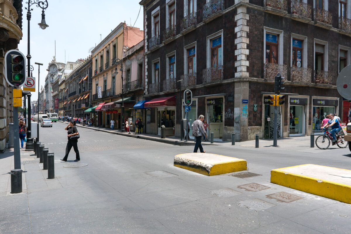 Intersection in Mexico City’s historic center with pedestrians crossing and cyclists moving along the street. Colonial-era buildings with balconies line the block, housing shops at street level. A bus and several storefronts with awnings are visible further down the road.