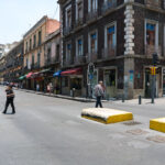 Intersection in Mexico City’s historic center with pedestrians crossing and cyclists moving along the street. Colonial-era buildings with balconies line the block, housing shops at street level. A bus and several storefronts with awnings are visible further down the road.