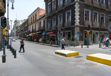 Intersection in Mexico City’s historic center with pedestrians crossing and cyclists moving along the street. Colonial-era buildings with balconies line the block, housing shops at street level. A bus and several storefronts with awnings are visible further down the road.