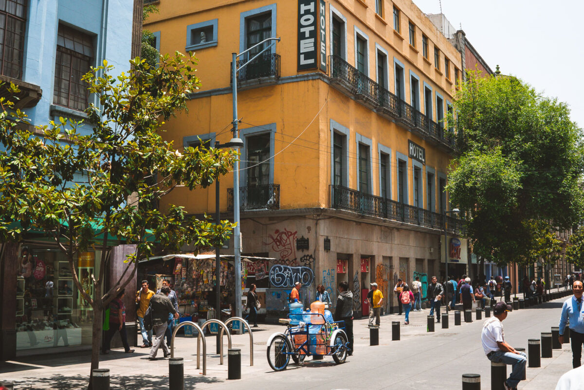 A street in Mexico City’s historic center with pedestrians walking past shops and vendors. A bright yellow hotel building with blue trim stands on the corner, while a tricycle cart carrying large water jugs moves through the scene. Trees line the walkway, and people gather along the shaded sidewalks.