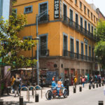A street in Mexico City’s historic center with pedestrians walking past shops and vendors. A bright yellow hotel building with blue trim stands on the corner, while a tricycle cart carrying large water jugs moves through the scene. Trees line the walkway, and people gather along the shaded sidewalks.
