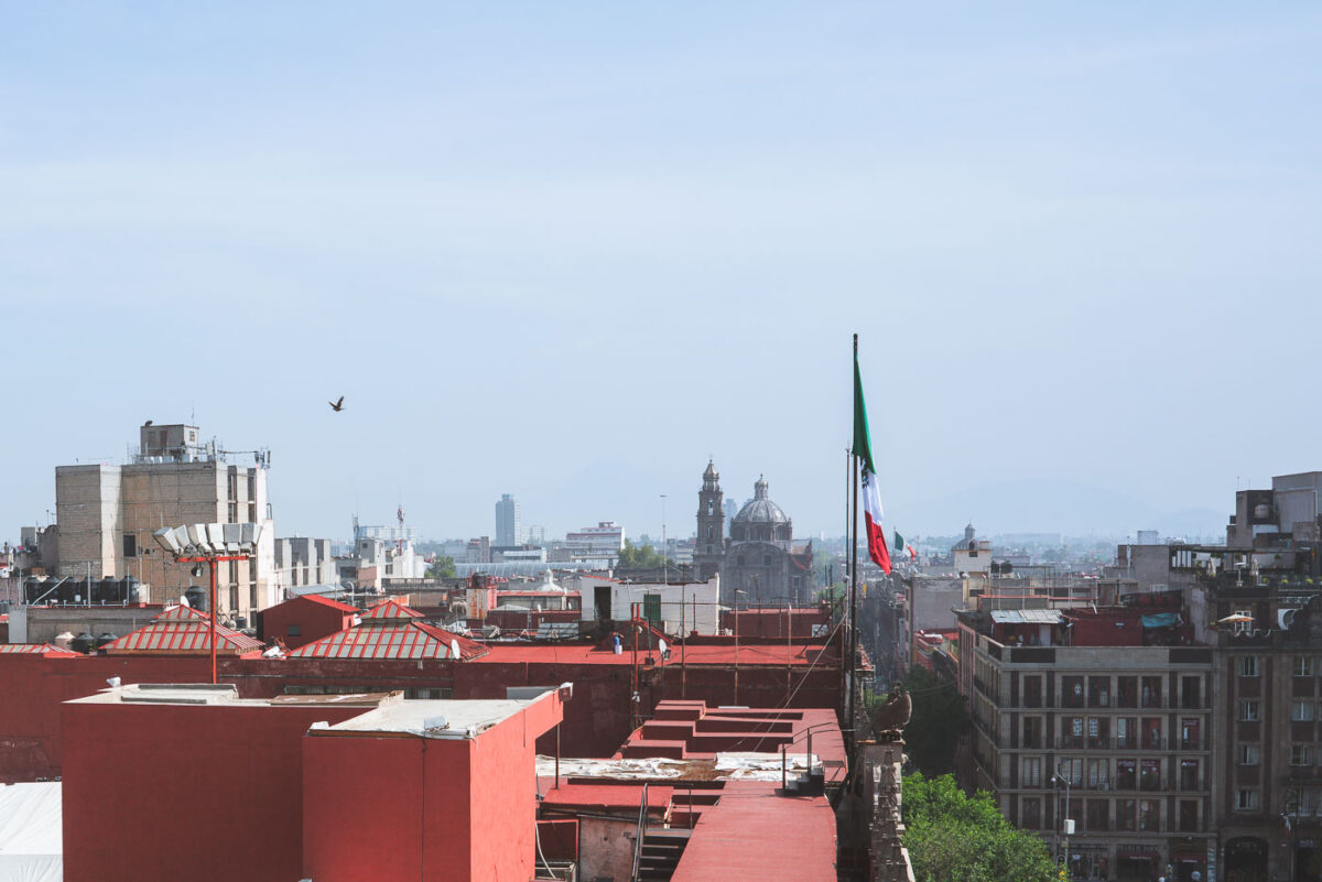 View over the rooftops of central Mexico City, where a large Mexican flag is raised above the buildings near the Zócalo. In the distance, church domes and towers rise over the historic district, with modern high-rises visible further out along the skyline.
