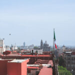 View over the rooftops of central Mexico City, where a large Mexican flag is raised above the buildings near the Zócalo. In the distance, church domes and towers rise over the historic district, with modern high-rises visible further out along the skyline.