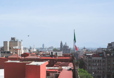 View over the rooftops of central Mexico City, where a large Mexican flag is raised above the buildings near the Zócalo. In the distance, church domes and towers rise over the historic district, with modern high-rises visible further out along the skyline.