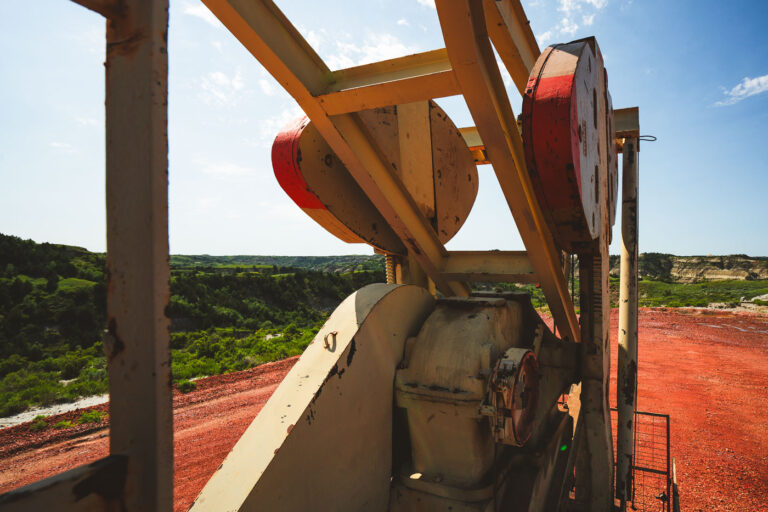Faided North Dakota Pumpjack 1 Viewed from behind the machinery, the pumpjack overlooks a sweeping stretch of North Dakota badlands. Its rusted gears and faded paint contrast sharply with the vivid red earth and green hills—a scene that captures both the rugged beauty and industrial imprint of the oil fields.