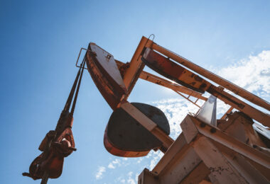Sunlight glints off the rusted steel of a working pumpjack, its rhythmic motion cutting through the quiet plains air. Shot from below, the towering machine looms against the sky—a reminder of the human industry that continues to shape the remote oil fields of the American West.