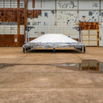 A completed dome section, likely part of the Space Launch System (SLS) core stage, sits wrapped and staged outside the Boeing production area at NASA’s Michoud Assembly Facility in New Orleans. The massive structure, used to cap propellant tanks, rests on a mobile transport frame against the weathered doors of an aging assembly hangar — a visual contrast between decades of industrial heritage and the cutting edge of modern rocketry. The surface reflections from the wet concrete emphasize the scale and symmetry of this aerospace component awaiting the next step in its journey toward integration.