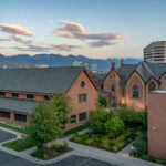 Evening light falls over downtown Salt Lake City, with the Wasatch Mountains silhouetted beyond. The historic Trinity Episcopal Cathedral, built of red sandstone in the Gothic Revival style, stands alongside modern offices and newer brick buildings. The contrast highlights the city’s mix of frontier-era architecture and contemporary growth at the edge of Utah’s mountain corridor.