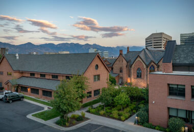 Evening light falls over downtown Salt Lake City, with the Wasatch Mountains silhouetted beyond. The historic Trinity Episcopal Cathedral, built of red sandstone in the Gothic Revival style, stands alongside modern offices and newer brick buildings. The contrast highlights the city’s mix of frontier-era architecture and contemporary growth at the edge of Utah’s mountain corridor.