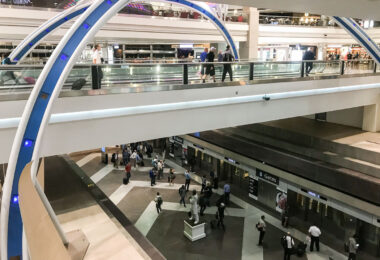 A walkway inside the Denver International Airport. Passengers wait below for the train to the B Gates as others roll luggage across elevated concourses. The scene captures the constant motion and layered architecture of one of the nation’s busiest air hubs.