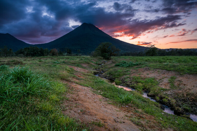 Arenal Volcano at sunset in Costa Rica, with dramatic clouds over the conical peak and a stream in the foreground.