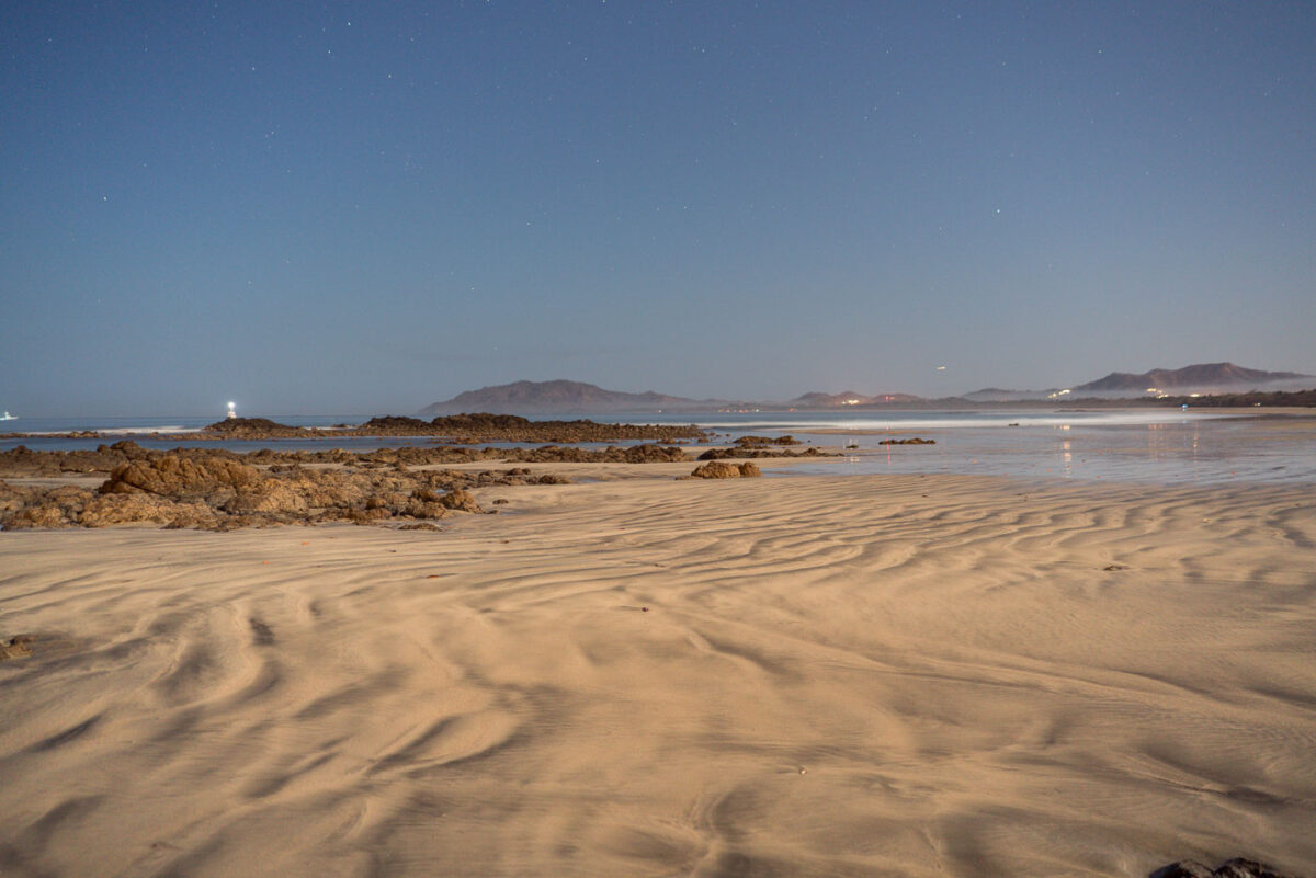 A lighthouse shines on a rocky coast at night, with distant lights along the shore and mountains in the background. Rippled sand and shallow water with rocks are in the foreground.