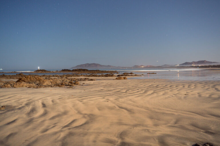 Night Beach with Lighthouse and Distant Lights 1 A lighthouse shines on a rocky coast at night, with distant lights along the shore and mountains in the background. Rippled sand and shallow water with rocks are in the foreground.