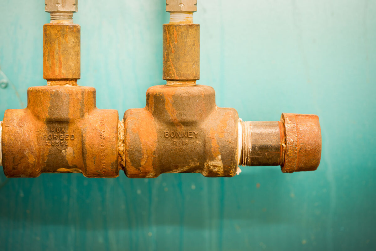 Rust-streaked industrial valves, stamped with “MOA Forged” and “BONNEY,” mounted against a teal wall, showing the wear of age and use.