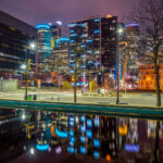 Downtown Minneapolis Reflections 2 Illuminated office and residential towers rise over downtown Minneapolis after dark, their façade lighting and interior floors forming a dense urban grid against a winter sky. The blue-accented crowns and vertical light bands highlight the city’s mix of late-20th-century corporate towers and newer high-rise residential development that has reshaped the downtown core since the 2000s. In the foreground, a still water feature mirrors the skyline, emphasizing the geometry and lighting strategies used to distinguish individual buildings within a tightly packed central business district. This area reflects Minneapolis’s broader downtown reinvestment, where office, residential, and public infrastructure coexist within a walkable, transit-served core.