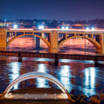 Raspberry Island Bandshell and Wabasha Street Bridge at Night 3 The Schubert Club Bandshell on Raspberry Island, illuminated along the Mississippi River in Saint Paul, Minnesota. Completed in 2002, the modern arched performance structure serves as an outdoor music venue with views toward the historic Wabasha Street Bridge, visible in the background. The bridge, originally built in 1859 and reconstructed in 1998, is known as the only true vertical align arch bridge over the Mississippi. Its decorative architectural lighting reflects across the water alongside the flooded riverbank, highlighting how the river’s seasonal water levels shape the island landscape. Raspberry Island has long served as a gathering space and riverfront access point, evolving from a railroad landing to a cultural park and performance venue.