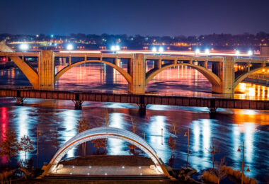 The Schubert Club Bandshell on Raspberry Island, illuminated along the Mississippi River in Saint Paul, Minnesota. Completed in 2002, the modern arched performance structure serves as an outdoor music venue with views toward the historic Wabasha Street Bridge, visible in the background. The bridge, originally built in 1859 and reconstructed in 1998, is known as the only true vertical align arch bridge over the Mississippi. Its decorative architectural lighting reflects across the water alongside the flooded riverbank, highlighting how the river’s seasonal water levels shape the island landscape. Raspberry Island has long served as a gathering space and riverfront access point, evolving from a railroad landing to a cultural park and performance venue.