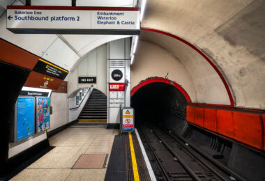 The southbound Bakerloo line platform at Charing Cross Underground Station in central London, showing the distinctive narrow tube tunnel built in the early 1900s. The red-edged lining around the tunnel mouth and the tiled passageway reflect classic London Transport design standards. Opened in 1906, the Bakerloo line was one of the early deep-level “tube” lines, and this station once served as its southern terminus before extensions carried it to Elephant & Castle. The signage and steep staircases illustrate the compact engineering that defined London’s early subterranean railways.