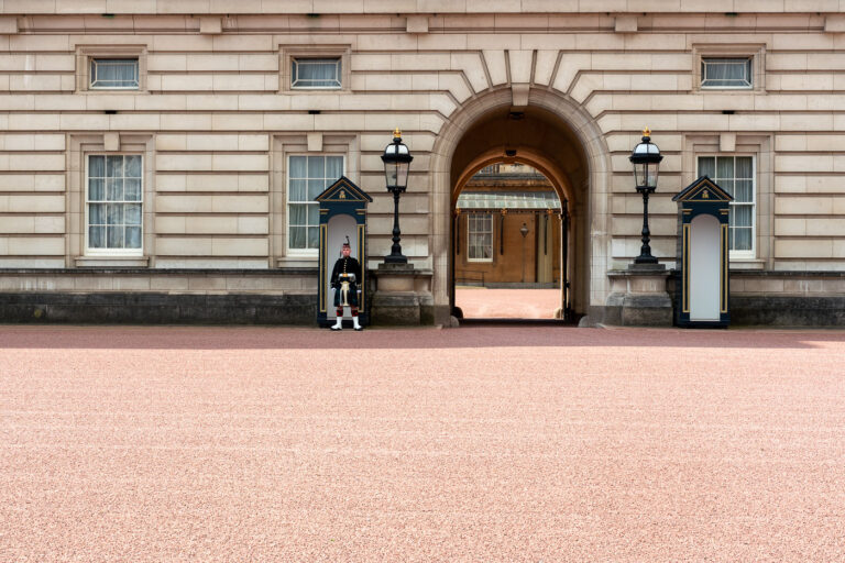 Guard at Buckingham Palace Archway 3 A member of the Queen’s Guard stands watch outside the archway of Buckingham Palace, London. The sentry is dressed in the traditional dark uniform with white belt and bearskin cap, symbolizing centuries of ceremonial military service. The arched passageway behind him connects the forecourt to the inner courtyard of the palace, the official residence of the British monarch since 1837. These guards belong to one of the Foot Guards regiments, trained soldiers who perform both ceremonial duties and active service in the British Army.