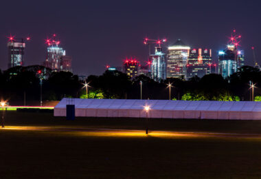 The illuminated towers of Canary Wharf rise beyond the trees of Greenwich Park, a contrast between London’s financial core and its historic green spaces. The red obstruction lights and construction cranes mark the district’s ongoing evolution, while the pyramid-topped One Canada Square and the glass towers of Citi and J.P. Morgan anchor the skyline. In the foreground, a temporary event structure sits quietly under soft amber streetlights, emphasizing the stillness of the park against the bright geometry of the city beyond.