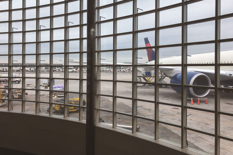 Delta Jet at Concourse A – Detroit Metro Airport 1 Viewed through the grid of Concourse A’s vast curtain-wall windows, a Delta Air Lines jet sits at the gate at Detroit Metro Airport, undergoing final preparations before departure. Ground crews maneuver baggage carts and service vehicles along the concrete apron beneath the overcast Michigan sky. The McNamara Terminal’s panoramic windows reveal the precision and coordination of modern aviation — a quiet balance of machinery, logistics, and architecture that defines one of America’s most important airline hubs.