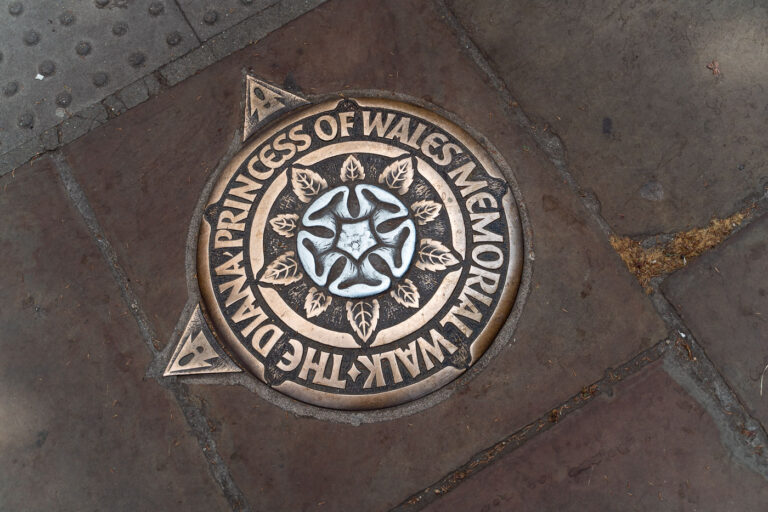The Diana Princess of Wales Memorial Walk Marker 1 Set into the pavements of central London, this bronze and aluminum marker denotes the Diana Princess of Wales Memorial Walk—a seven-mile circular route passing through four of the city’s royal parks: St. James’s Park, Green Park, Hyde Park, and Kensington Gardens. Installed in 2000, the trail honors the late Princess Diana’s humanitarian legacy, guiding walkers past many places associated with her life. Each plaque features an English rose at its center, surrounded by leaves and directional points that quietly lead thousands of visitors along the commemorative path each year.