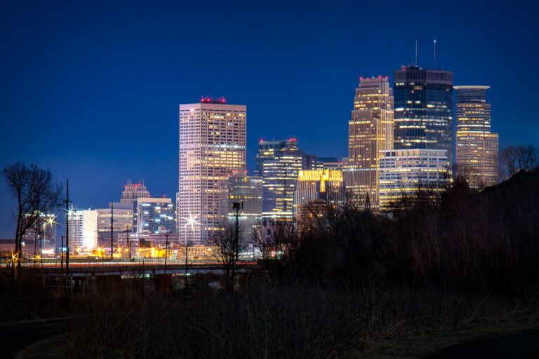 Downtown Minneapolis from Cedar Lake Trail 3 The Minneapolis skyline glows against a deep twilight sky, its mix of modern glass towers and classic architecture illuminated from within. Iconic buildings such as the IDS Center and Wells Fargo Center define the city’s vertical rhythm, while the rail and industrial foreground recall the infrastructure that shaped its early economy. Captured during blue hour, the scene reflects the enduring balance between industry and innovation that characterizes Minneapolis.