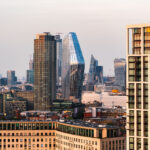 A view across central London at dusk, where residential towers and commercial high-rises stand against the soft evening light. Dominating the center is One Blackfriars, the glass-clad, sculptural tower completed in 2019, reflecting the sky with its curved façade. To its right is 22 Bishopsgate under construction, rising as one of the city’s tallest buildings. Further east, the distinctively slanted shape of The Scalpel and the concave form of 20 Fenchurch Street, commonly known as the Walkie-Talkie, mark the financial district’s development in the 2010s. The variety of architectural styles reflects London’s rapid vertical growth and economic shift toward high-density mixed-use urban living near the River Thames.
