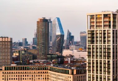 A view across central London at dusk, where residential towers and commercial high-rises stand against the soft evening light. Dominating the center is One Blackfriars, the glass-clad, sculptural tower completed in 2019, reflecting the sky with its curved façade. To its right is 22 Bishopsgate under construction, rising as one of the city’s tallest buildings. Further east, the distinctively slanted shape of The Scalpel and the concave form of 20 Fenchurch Street, commonly known as the Walkie-Talkie, mark the financial district’s development in the 2010s. The variety of architectural styles reflects London’s rapid vertical growth and economic shift toward high-density mixed-use urban living near the River Thames.