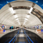 Commuters ride the long escalators inside Charing Cross Underground Station in central London. The station, a busy interchange on the Northern and Bakerloo lines, is known for its deep tunnels, heavy foot traffic, and role as a gateway to Trafalgar Square and the West End. Rows of posters line the walls, while bright fluorescent lighting and metallic finishes highlight the stark, utilitarian design typical of the London Tube.