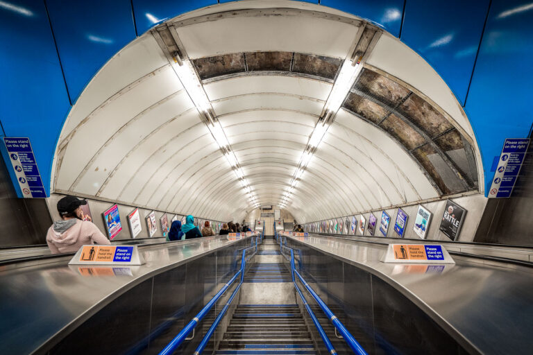 Charing Cross Escalator in London 3 Commuters ride the long escalators inside Charing Cross Underground Station in central London. The station, a busy interchange on the Northern and Bakerloo lines, is known for its deep tunnels, heavy foot traffic, and role as a gateway to Trafalgar Square and the West End. Rows of posters line the walls, while bright fluorescent lighting and metallic finishes highlight the stark, utilitarian design typical of the London Tube.