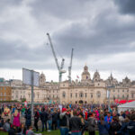 Participants and supporters gather at Horse Guards Parade near the finish area of the 2019 London Marathon. The scene captures the mix of exhaustion and celebration as runners cross the line against a backdrop of historic architecture and temporary infrastructure erected for the event. Cranes tower above the domes and facades of Whitehall’s government buildings, underscoring the juxtaposition of London’s centuries-old civic landmarks and its ever-changing skyline. The annual marathon, one of the world’s largest, draws tens of thousands of athletes and spectators to central London, filling the ceremonial grounds with color, energy, and determination.