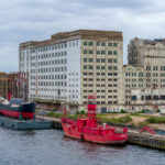 Once a centerpiece of London’s industrial age, Millennium Mills stands derelict on the south side of the Royal Victoria Dock in Silvertown. Built in 1905 for Spillers, the flour mill operated for decades before closing in the 1980s. The weathered façade and faded signage remain prominent features of the docklands skyline. Moored in front is the striking red Trinity House Lightship 93, formerly used as a floating lighthouse to warn ships at sea. Today, the area is part of the Royal Docks regeneration zone, blending remnants of Britain’s maritime and industrial heritage with modern redevelopment.