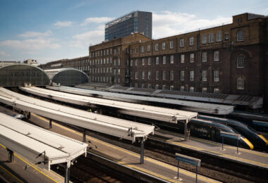An elevated view of London Paddington Station shows the sweeping iron-and-glass train sheds designed in the mid-19th century for the Great Western Railway by Isambard Kingdom Brunel. The long canopies stretch over multiple platforms, where modern GWR Class 800 “Intercity Express” trains await departure. The adjacent red-brick offices of the original railway company still stand beside the terminus, a reminder of Paddington’s role as both a Victorian engineering triumph and a key link in Britain’s modern high-speed network.